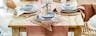 Gedekte brunchtafel, eetkamermeubels van natuurlijk hout en rotanweefsel, keramisch servies, roze glazen en goudkleurig bestek op een tafelloper van katoen.