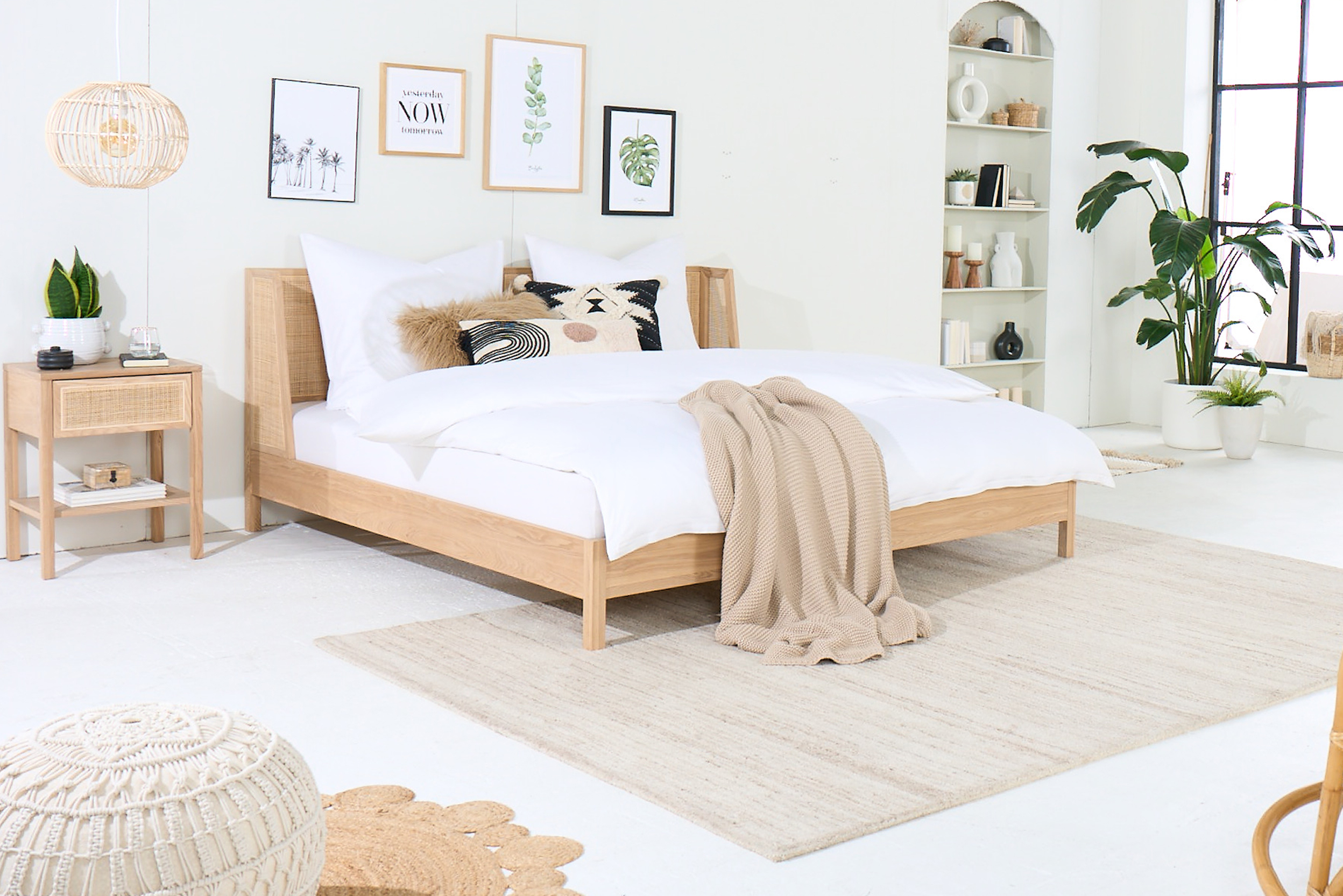 Schlafzimmer mit hellen Holzmöbeln der Serie Taylor by kollected, einer Exklusivmarke von home24: ein Bett, ein Nachttisch, ein Schaukelstuhl, ein Schreibtisch inkl. Schreibtischstuhl, dazu viele Grünpflanzen, Teppiche, Textilien und ein Strickpouf. Allen Taylor-Produkten gemein ist die Kombination aus hellem Eichenholz und Rattan.