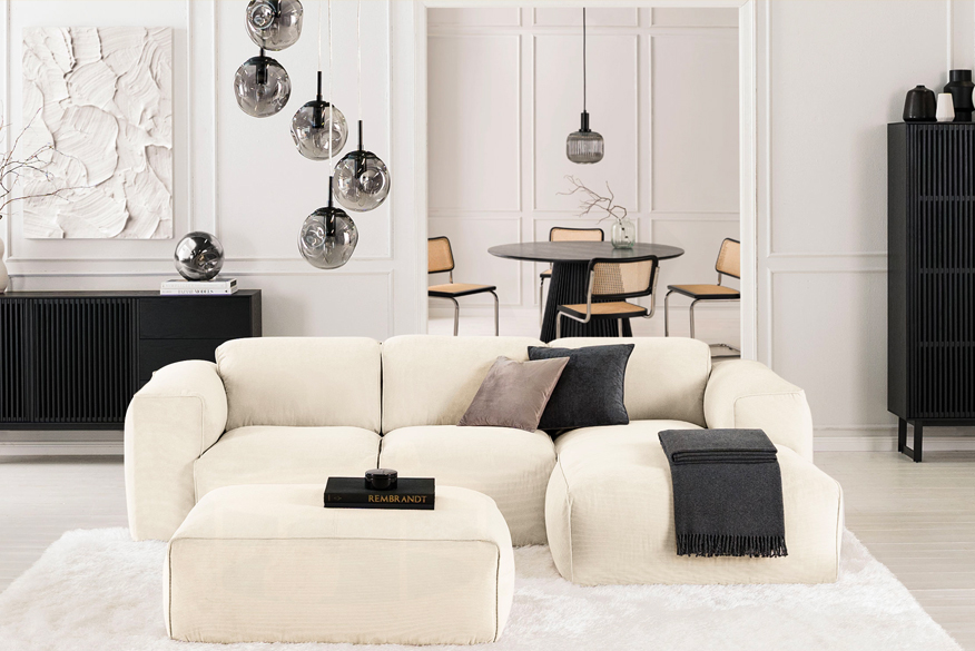 Wohnzimmer mit beigem Cordsofa und Cordhocker der Serie Hudson by Studio Copenhagen sowie Sideboard, Schrank und Esstisch der Serie Qara by Studio Copenhagen in Schwarz; dazu Pendelleuchten aus grauem Rauchglas, Flechtstühle und ein Servierwagen.