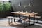 Patio mit Großstadtflair: Outdoor-Möbel aus lackiertem Metall und massivem Akazienholz auf einer Terrasse aus gegossenem Beton, kombiniert mit Industrieleuchten und funktionaler Deko, daneben ein Pflanztopf mit Ziergräsern.