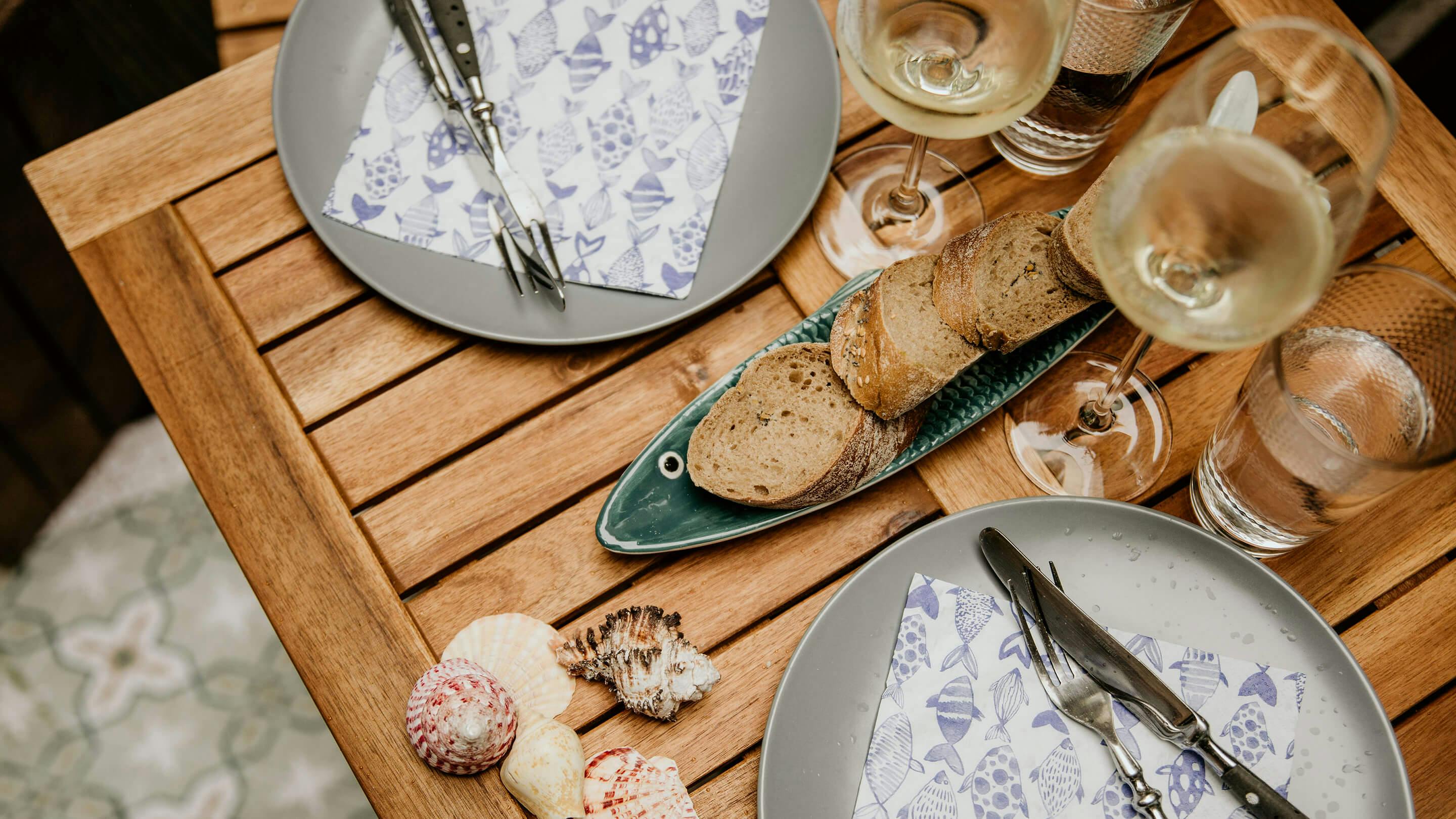 Gedeckter Gartentisch von oben gesehen: Teller mit Besteck und Serviette, Baguette in Scheiben, Weingläser (Marke: Butlers).