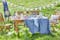 Gartenmöbelset aus Holz von BUTLERS, platziert auf einem grünen Rasen für ein Picknick: dazu Geschirr, Gläser und eine Etagere sowie ein weißes Kissen und eine blaue Tischdecke.