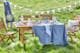 Gartenmöbelset aus Holz von BUTLERS, platziert auf einem grünen Rasen für ein Picknick: dazu Geschirr, Gläser und eine Etagere sowie eine weißes Kissen und eine blaue Tischdecke.