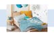 Helles Kinderbett aus Holz, auf dem Bettwäsche und Plaid in Ocker, Blau und Steingrau drapiert sind; daneben ein beiger Kinderteppich mit Fransen, auf dem ein getuftetes Kissen mit Regenbogen sowie Spielsachen liegen.