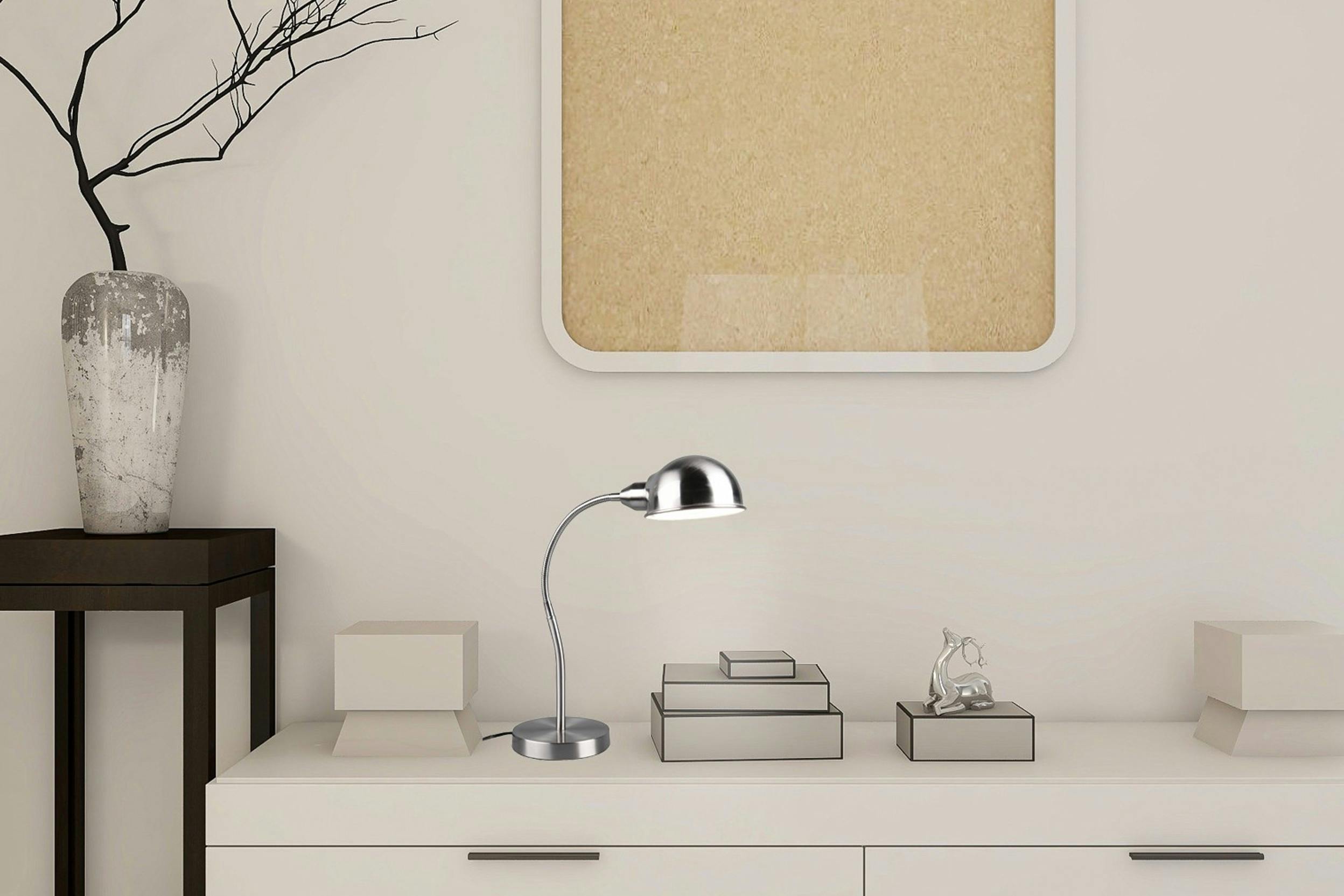 Lampe de bureau argentée posée sur une enfilade blanche, entourée d'accessoires décoratifs, devant un mur beige.