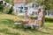 Collage met twee foto's: links een zonnige, groene tuin met klassiek houten tuinmeubels, witte slingers boven de tafel en twee vrouwen die de tafel dekken; rechts een houten bankje met daarop een blauwgrijs kussen en deken, een koffiekopje en daaronder een lichtbruine hond die op kiezels ligt.