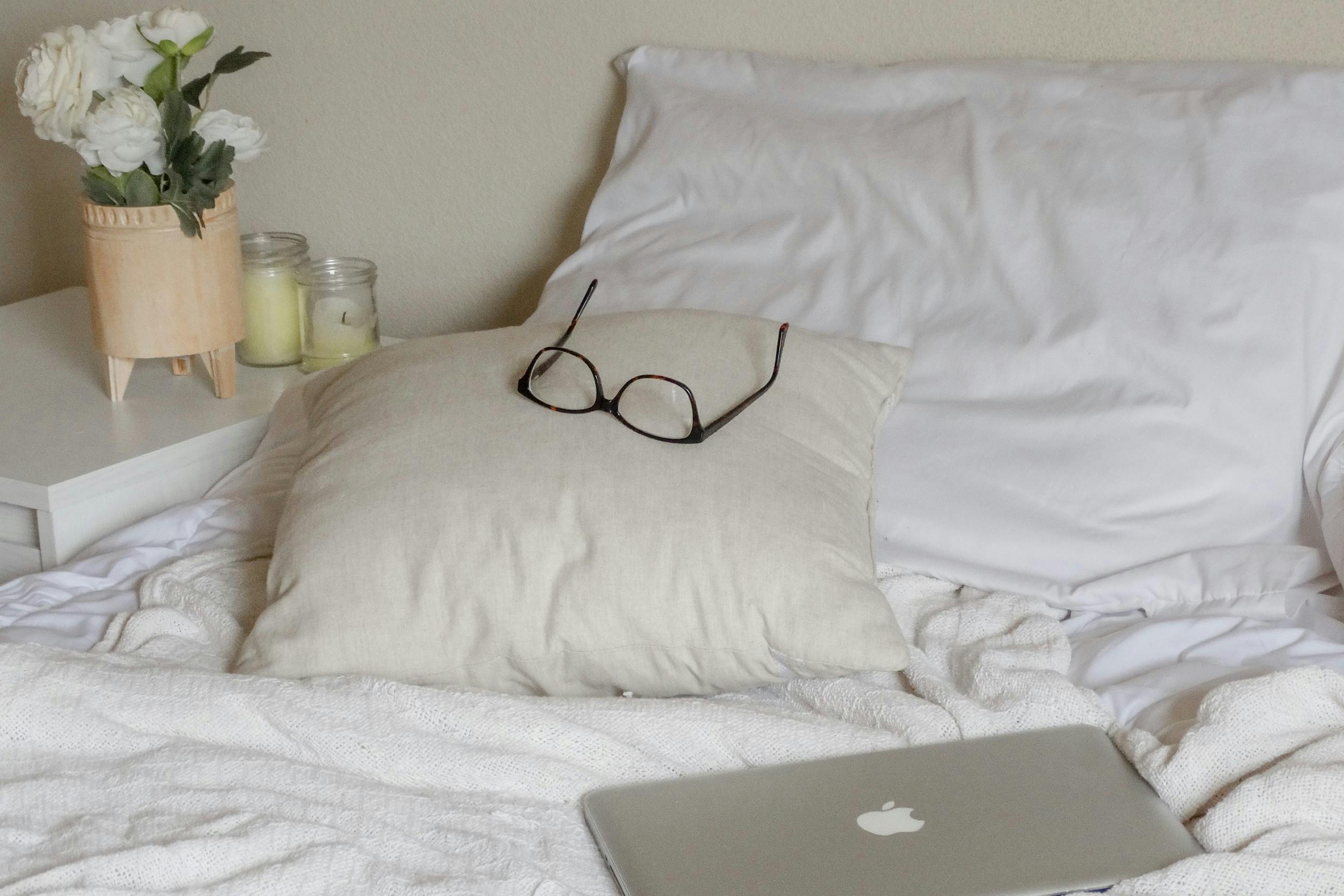 Bett mit heller Bettwäsche, schwarzer Lesebrille und einem Laptop