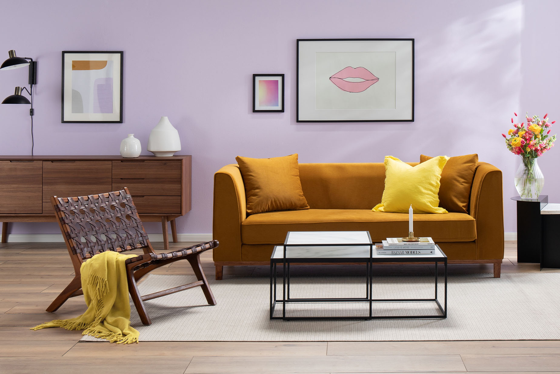 Wohnzimmer mit fliederfarbener Wand und Bildern, dazu ein senfgelbes Sofa mit gelbem Kissen, ein Couchtisch aus schwarzem Metall, ein brauner Ledersessel in Flechtoptik sowie ein Retro-Sessel mit grauem Polster und Holzbeinen.