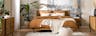 Schlafzimmer im Boho-Stil mit Palmentapete, Möbeln der home24 Marke kollected, braunen und weißen Textilien, Strickpouf, Teppich, Rattanlampe, einem schwarzen Spiegel und schwarzen Vasen.