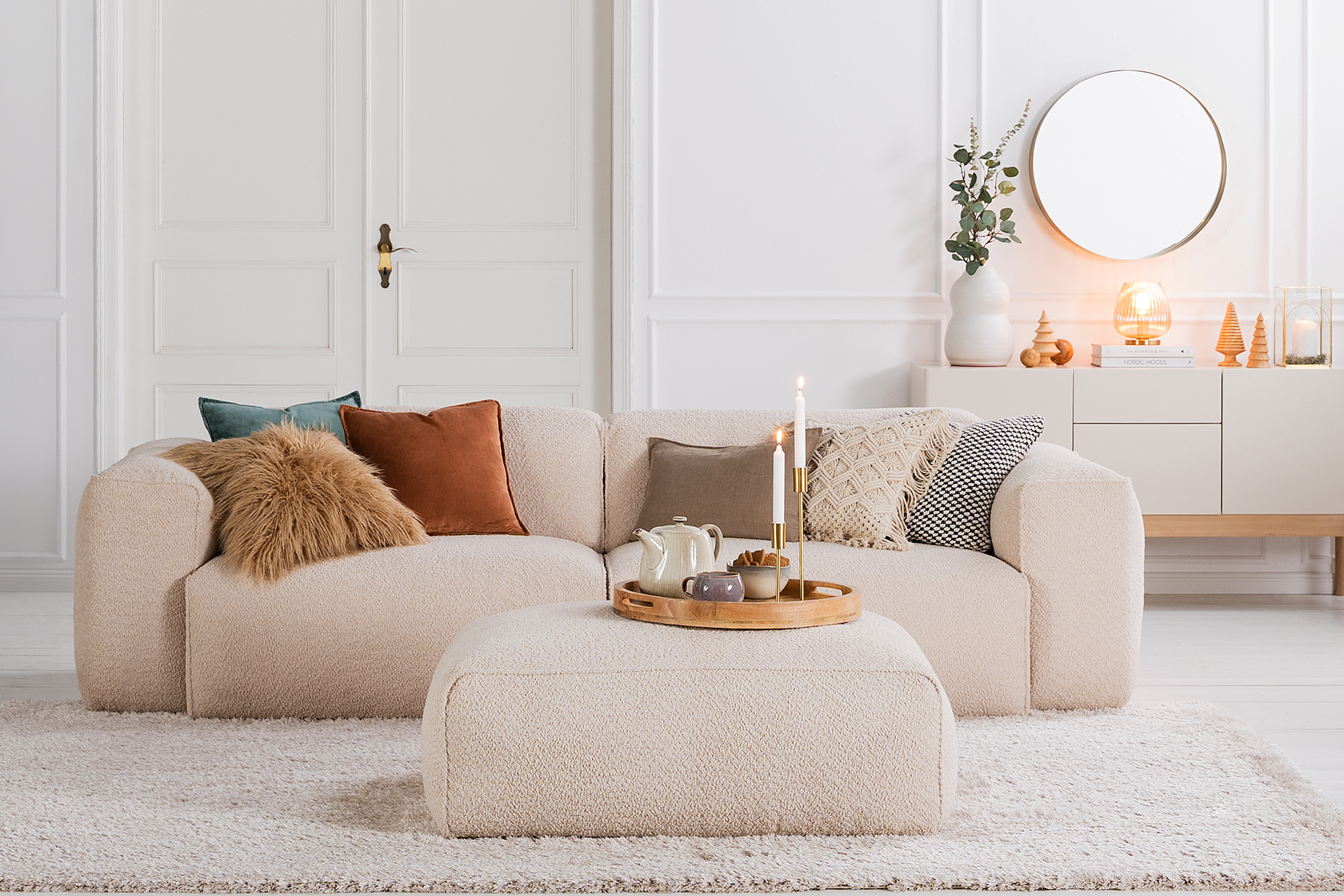Wohnzimmer mit Skandi-Möbeln der home24 Exklusivmarke Studio Copenhagen, wie zum Beispiel das beige Bouclésofa Hudson, Sessel Garbo oder Sideboard Cooby I. Dazu weihnachtliche Deko, Weihnachtsbäume, Kissen, Teppich und Plaids in Erdtönen, ein goldener Spiegel, Vasen und Tischleuchten.