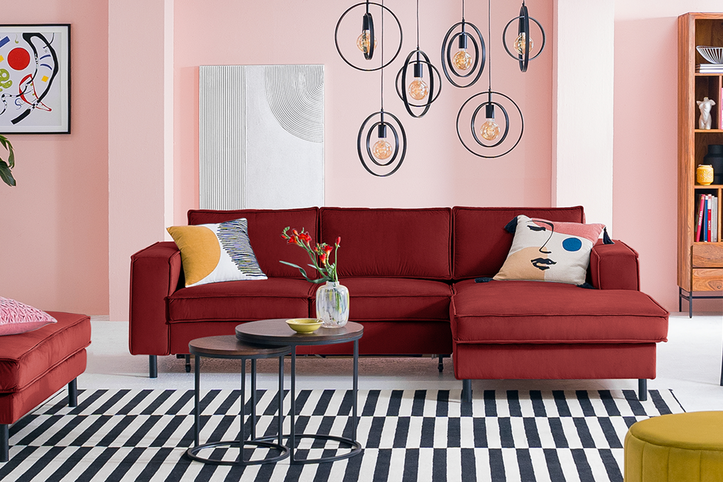Rotes Sofa inkl. Polsterhocker der home24 Exklusivserie Fort Dodge by kollected mit bunten Kissen auf einem schwarz-weiß gestreiften Teppich. Dazu zwei runde Beistelltische aus dunklem Holz und Metall im Industrial-Stil, ein gelber Pouf, ein schwarzer Stuhl mit Wiener Geflecht und gelbem Kissen, mehrere schwarze Pendelleuchten, ein Kandinsky-Wandbild und Highboard Deva aus Holz und Metall.