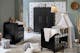 Chambre bébé avec meubles en bois noir et mur taupe : lit à barreaux avec linge de lit beige et blanc, commode à langer avec peluches, tapis beige, coussins clairs et suspension en rotin