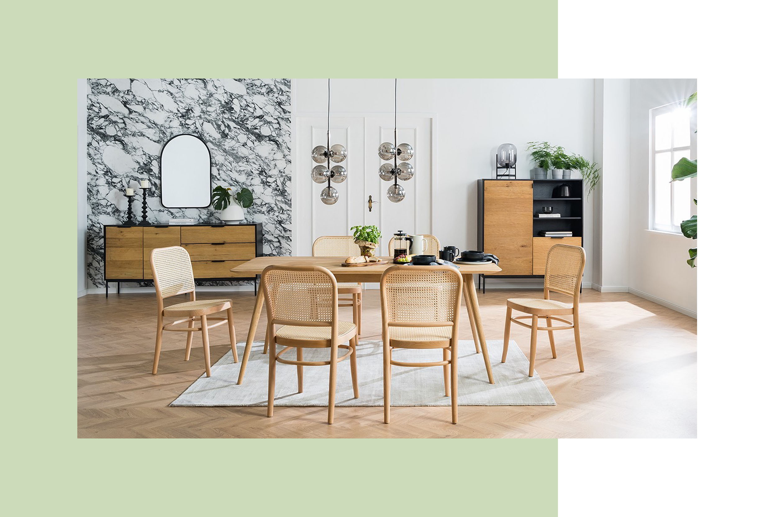 Heller Holztisch mit sechs Esszimmerstühlen mit Wiener Geflecht in einem Altbau-Esszimmer mit Holzboden, weißen Wänden, Kugellampen und Kommoden im Hintergrund, wobei die linke vor einer schwarz-weiß marmorierten Tapete steht