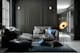 Grande pièce aux murs noirs avec grandes fenêtres, meubles gris foncés, coussins bleus, tapis gris et lampes noires