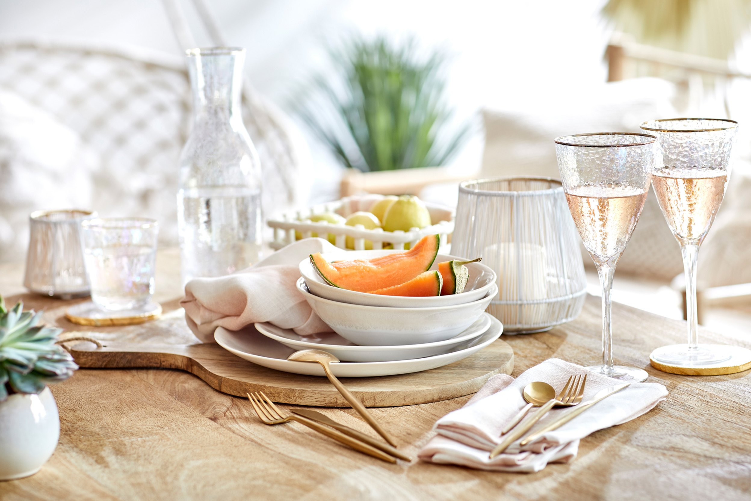 Esstisch aus Holz gedeckt mit weißem Geschirr und stilvollen Gläsern mit Goldrand, dazu goldfarbenes Besteck.