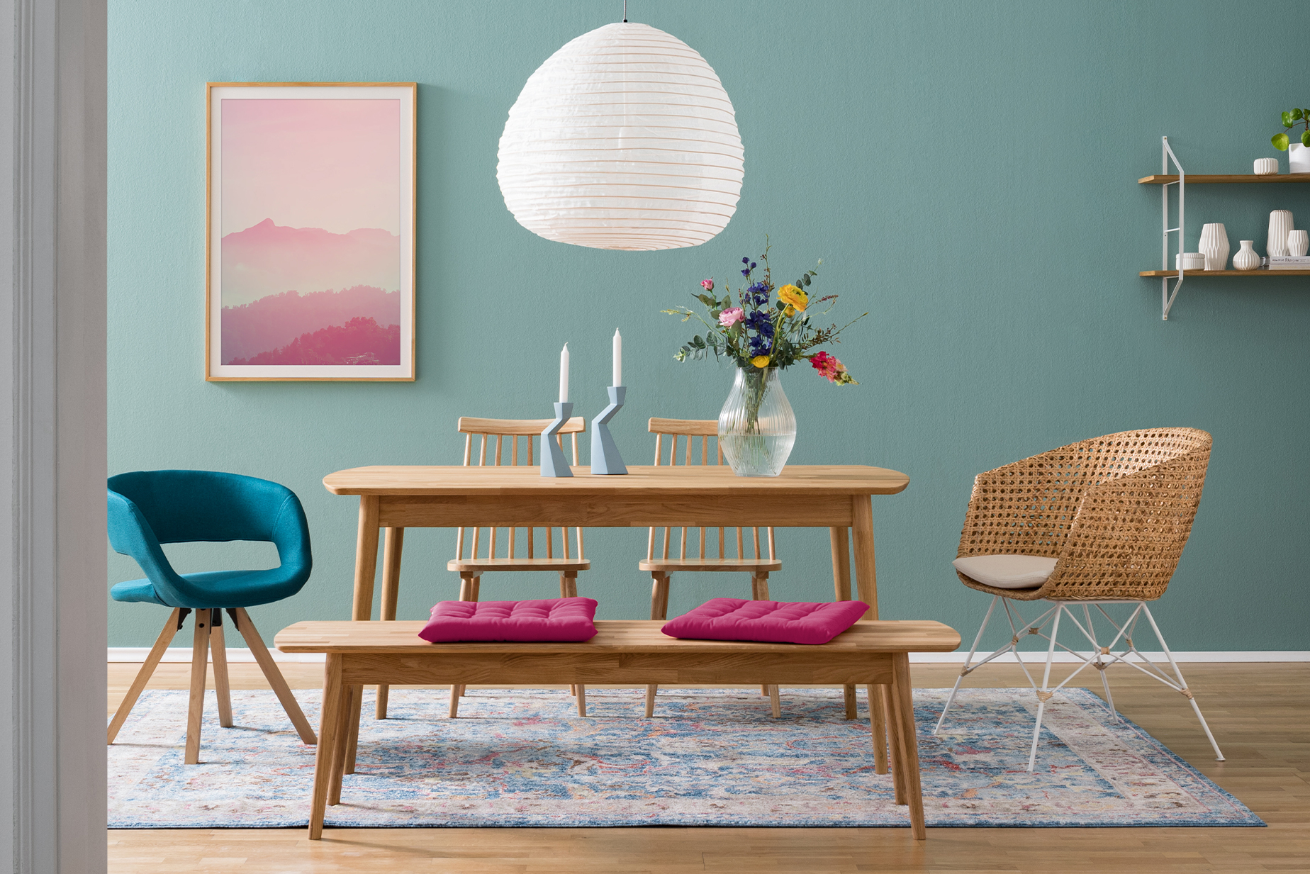Esszimmer in Blau- und Rosatönen mit einem Esstisch und einer Küchenbank aus hellem Massivholz und pinkfarbenen Sitzkissen, dazu Deko, Holzstühle, ein blauer Polsterstuhl sowie ein Esszimmerstuhl aus Rattan vor einer blauen Wand mit Bild und Wandregal