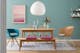 Eetkamer in blauwe en roze tinten met een eettafel en een keukenbank van licht massief hout en roze zitkussens, plus decoratie, houten stoelen, een blauwe gestoffeerde stoel en een eetkamerstoel van rotan voor een blauwe muur met foto en wandplank
