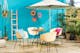 Terras met turquoise muur met een tuintafel in de industriële stijl, lichtgele kuipstoelen, een beige gebreide poef, een witte parasol, een kleurrijke hangmat en muurdecoratie, plus veel planten in bloempotten en hangende manden.
