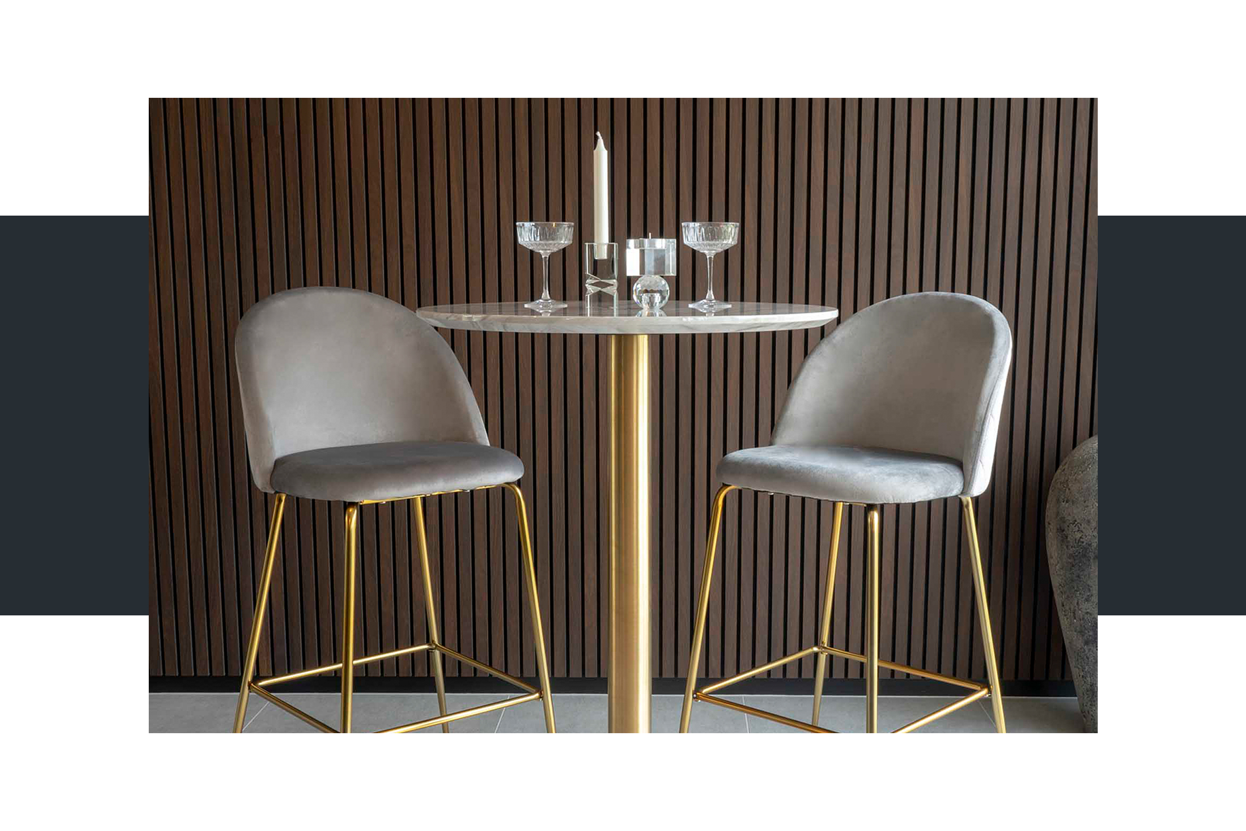 Bartisch mit weißer Marmorplatte und goldenem Tischbein, auf dem Champagnergläser und Kerzenständer stehen, sowie gepolsterte Barstühle aus hellgrauem Samt mit Goldbeinen; daneben ein goldener Servierwagen mit Spiritousen und Gläsern.