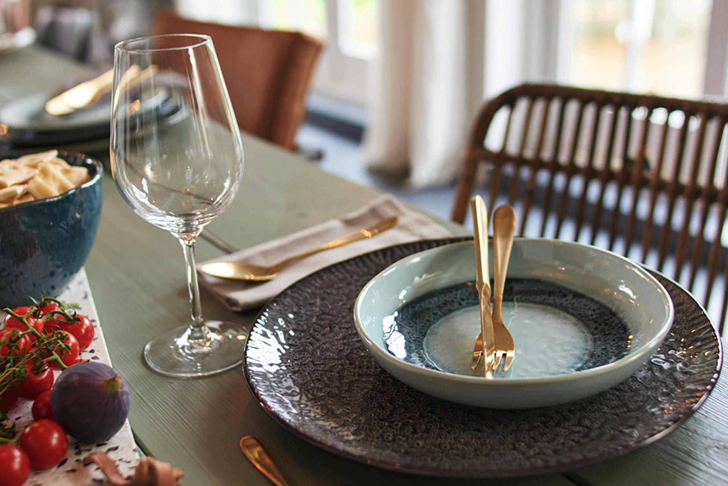 Gedekte tafel met dinerbord, soepbord en goudkleurig bestek, wijnglas, tomaten.