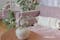 Lilafarbenes Sofa mit weißen Kissen im Boho-Style, dazu viele Grünpflanzen; daneben eine Glasvase mit rosa Pfingstrosen, dahinter eine Blumentapete.