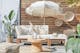 Loungemeubels voor buiten uit de LEXI by BUTLERS-serie van licht acaciahout met witte kussens, een bijpassende tafel versierd met witte vazen, een witte parasol met franjes, een outdoor-vloerkleed en poef van jute, evenals wanddecoratie, een plantenmand en lantaarn met gevlochten look.