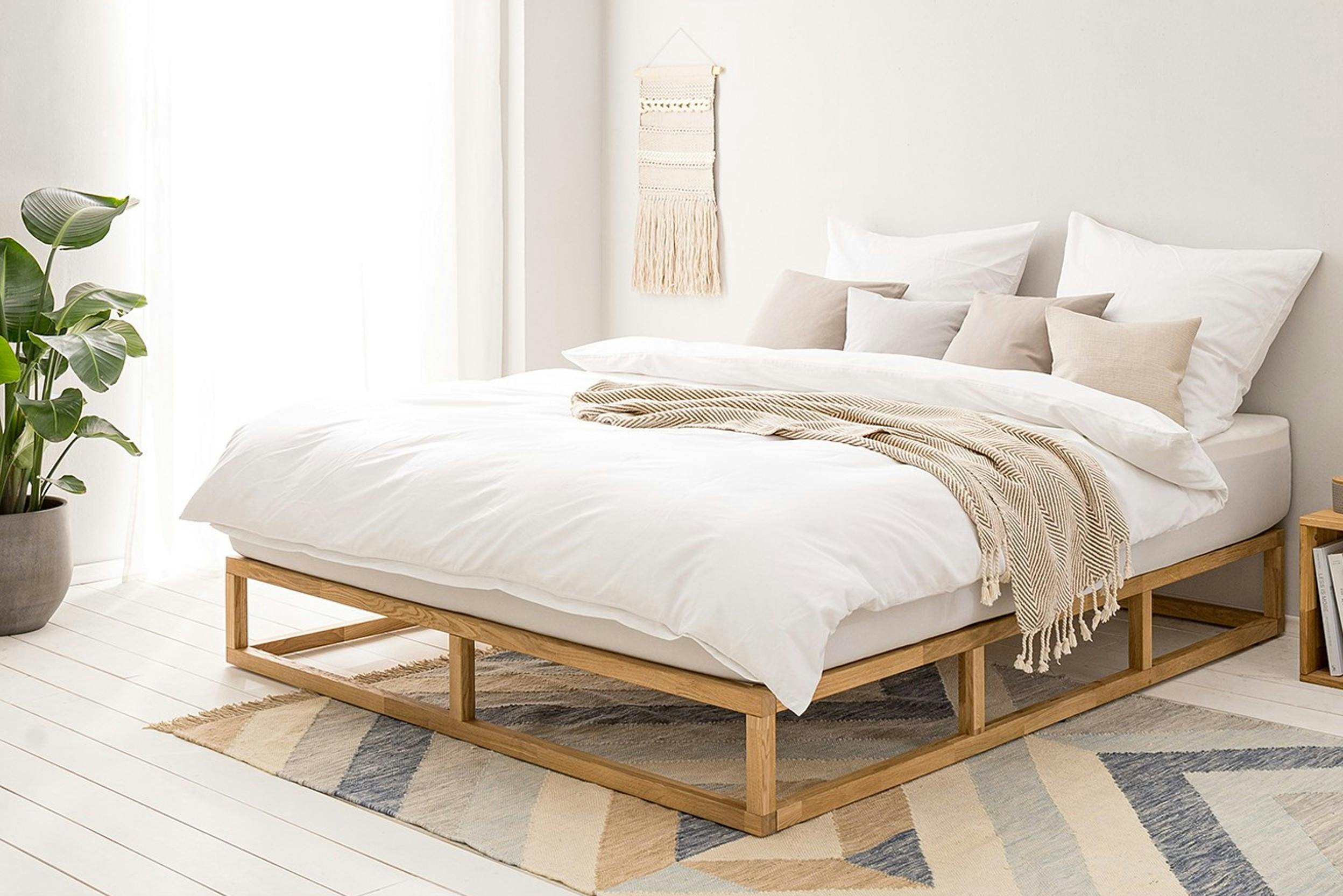 Helles Schlafzimmer mit Bett mit heller Bettwäsche, Teppich, Pflanze und Makramee-Wandbehang