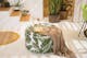 Pouf mit Palmenmotiven, dazu eine braune Decke, eine Karaffe und Gläser sowie ein Outdoor-Teppich, im Hintergrund viele Pflanzen in Blumentöpfen und Körben.