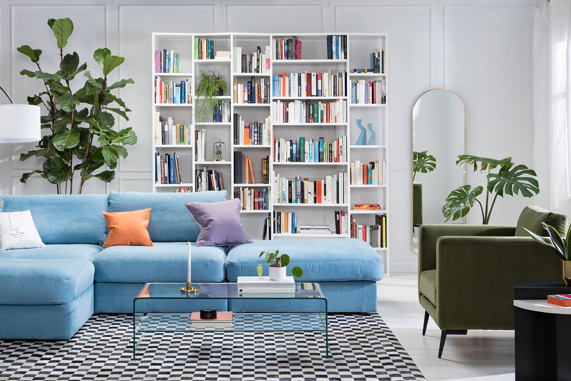 Wohnzimmer mit hellblauem Sofa, bunten Kissen, einem Glastisch sowie einem grünem Samtsessel, dazu eine Bogenlampe mit weißem Schirm, ein weißes Regal und ein schwarz-weiß karierter Teppich.