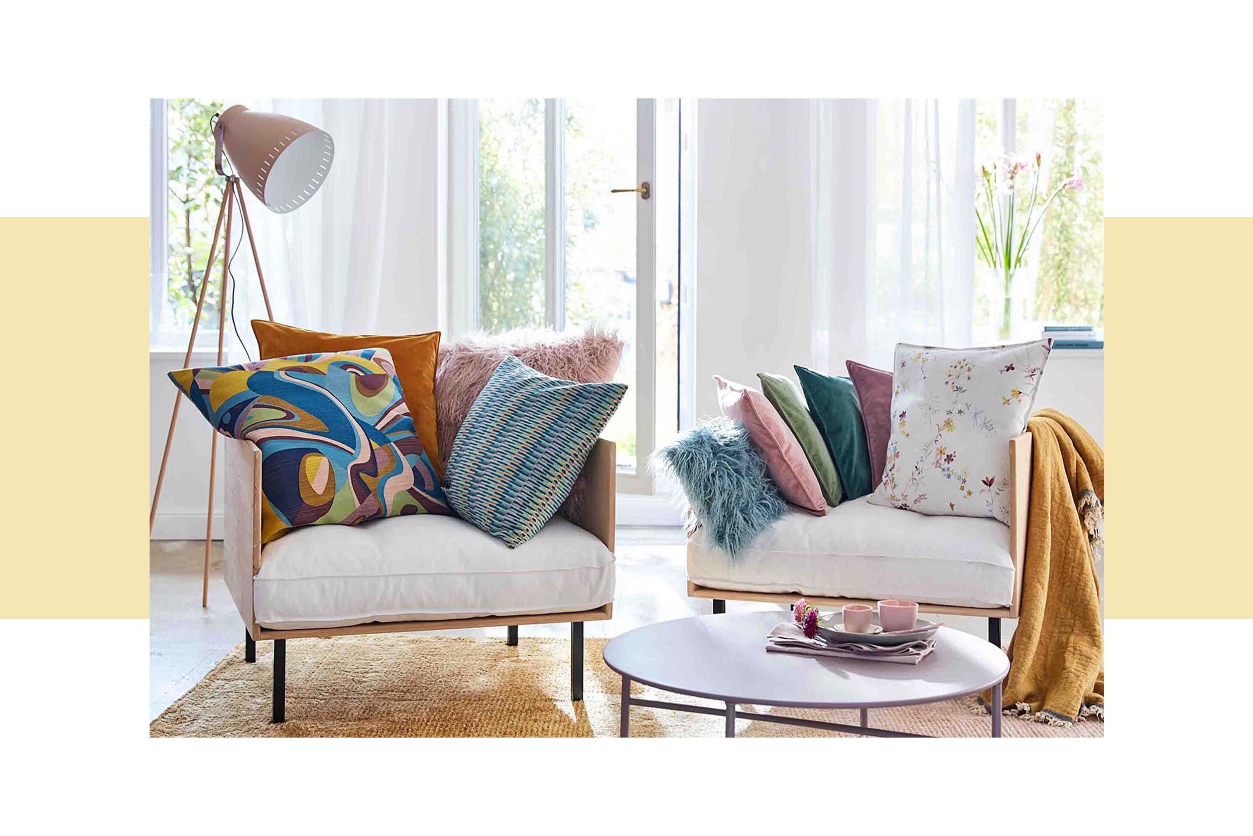 Zwei stylishe Sessel in Beige mit bunten Kissen in verschiedenen frühlingshaften Mustern; daneben sieht man auf einen bunten Teppich und eine Vase mit einem rosanen Blumenstrauß 