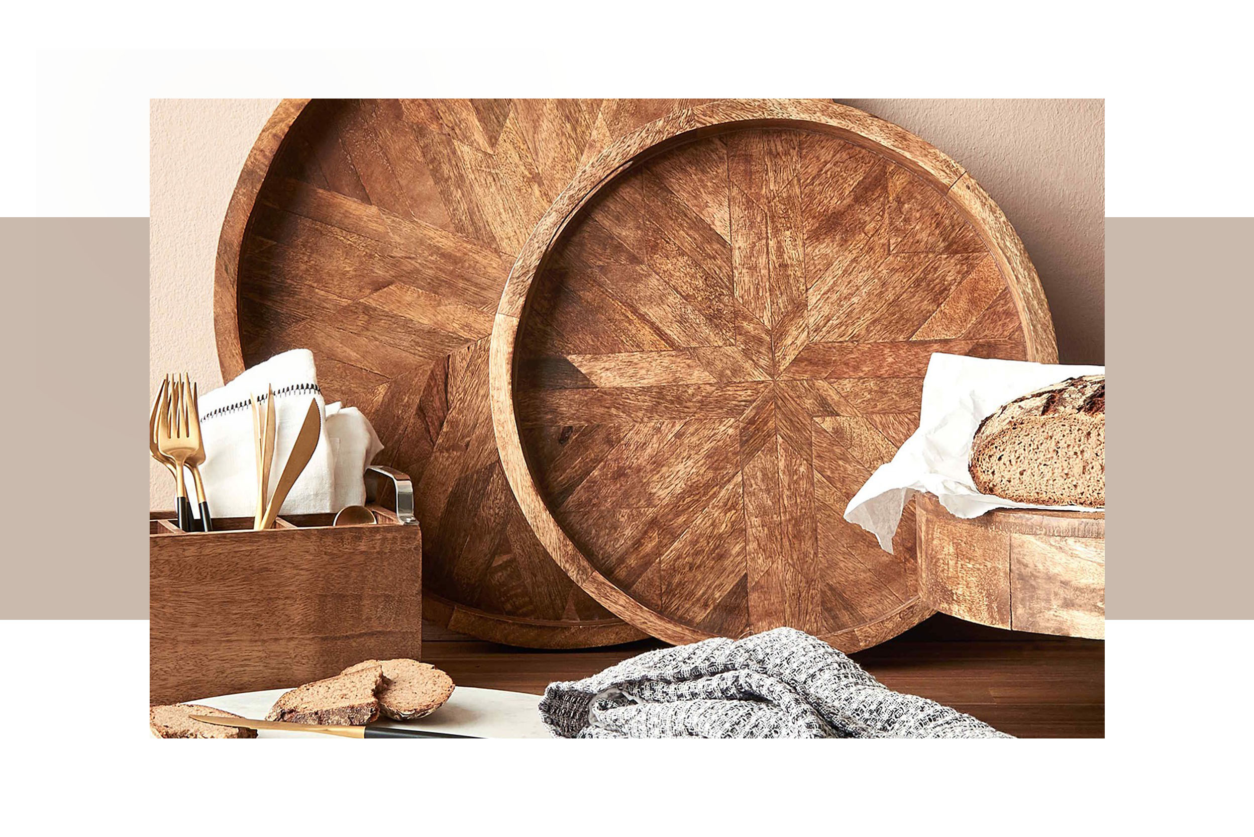 Zwei unterschiedlich große, runde Tabletts aus Mangoholz sowie ein Schneidebrett und Besteckkasten aus Holz mit goldenem Besteck und grauem Küchentuch davor liegend; daneben eine Ansammlung von weißen Vorratsbehältern der BUTLERS-Kollektion KARLTON BROS mit Holzdeckel und schwarzer Beschriftung 