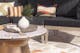 Bronzefarbener Gartentisch mit Terrazzo-Platte vor einem grauen Loungesofa mit gemusterten Deko-Kissen, dazu eine Deko-Platte mit weißer Vase.