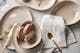 Auf einem Tisch mit cremefarbener Leinentischdecke steht beiges Geschirr in organischen Formen sowie Brot, goldenes Besteck und passende Leinenservietten