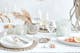 Weißer Tisch gedeckt mit Weingläsern, Windlichtern aus Glas, Untersetzern mit Muscheln, weißen Tellern und Schalen sowie einer silbernen Deko-Vase in Schneckenoptik.