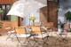 Kiesbett-Garten mit Tisch und Gartenstühlen im eleganten Landhausstil aus schwarzem Metall und Akazienholz, dazu weiße Sitzkissen, ein weißer Sonnenschirm, Grünpflanzen und eine Blumenvase aus Glas.