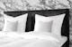 Boxspring KINX in witte structuurstof met wit beddengoed en een grijze deken, op de achtergrond een muur in marmeren look. Daarnaast twee zwart-witte hanglampen, een wit pluizig vloerkleed en meubels in de retrostijl van zwart hout met Weens vlechtwerk.