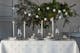 Festtagstafel mit weihnachtlicher Tischdecke und Kissen in Weiß und mit silbernen Sternen, dazu vier hohe silberne Kerzen, weißes Geschirr, eine graue Dekoschale, graue dicke Kerzen, eine Sternenkette mit Tannendeko sowie eine Lichterkette mit Glaskugeln, außerdem weiß-glänzende Weihnachtskugeln auf den Tellern.