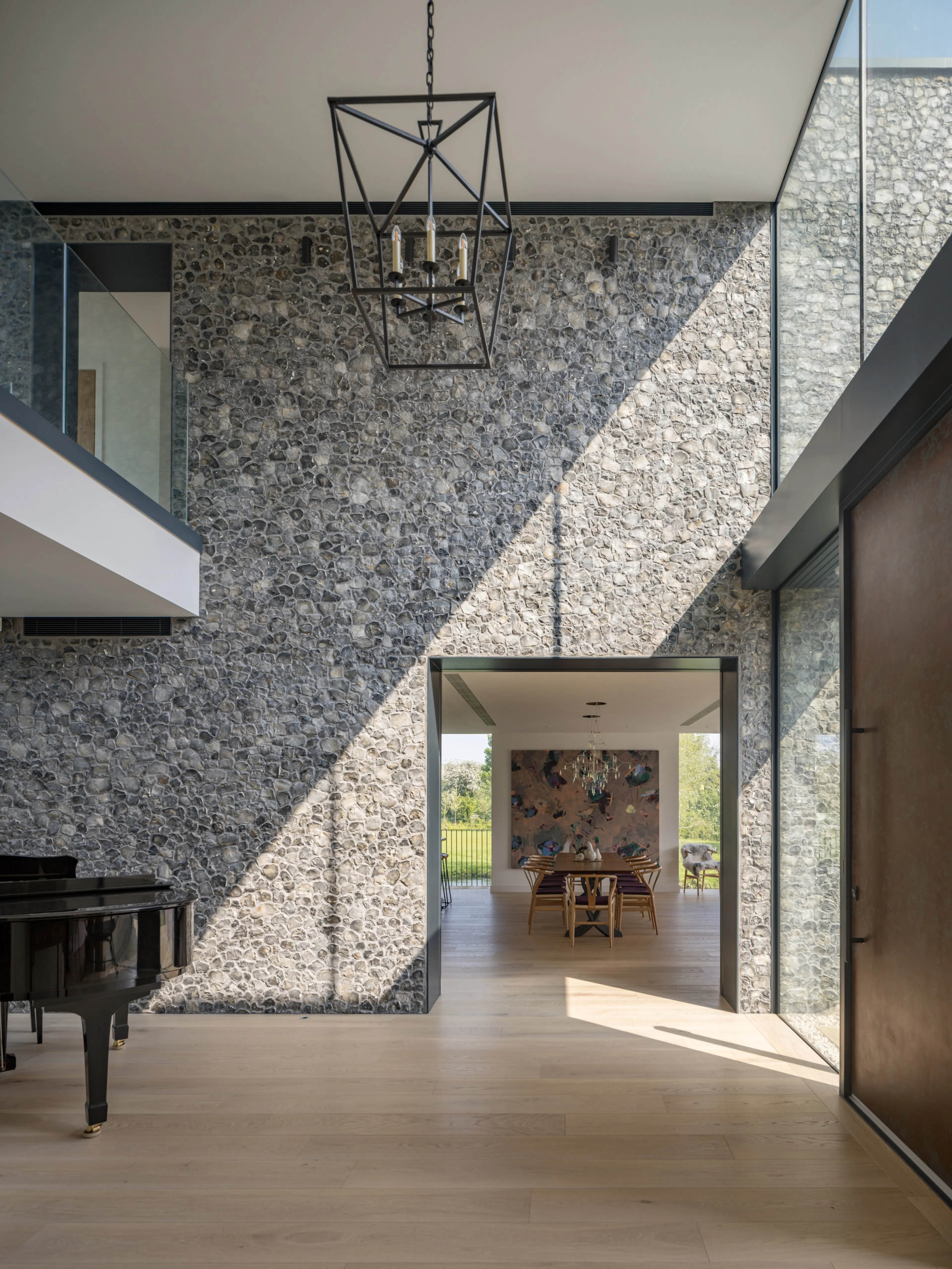 Double-height hall with a grand piano against a flint wall, sunlight falling across it, and the dining room beyond