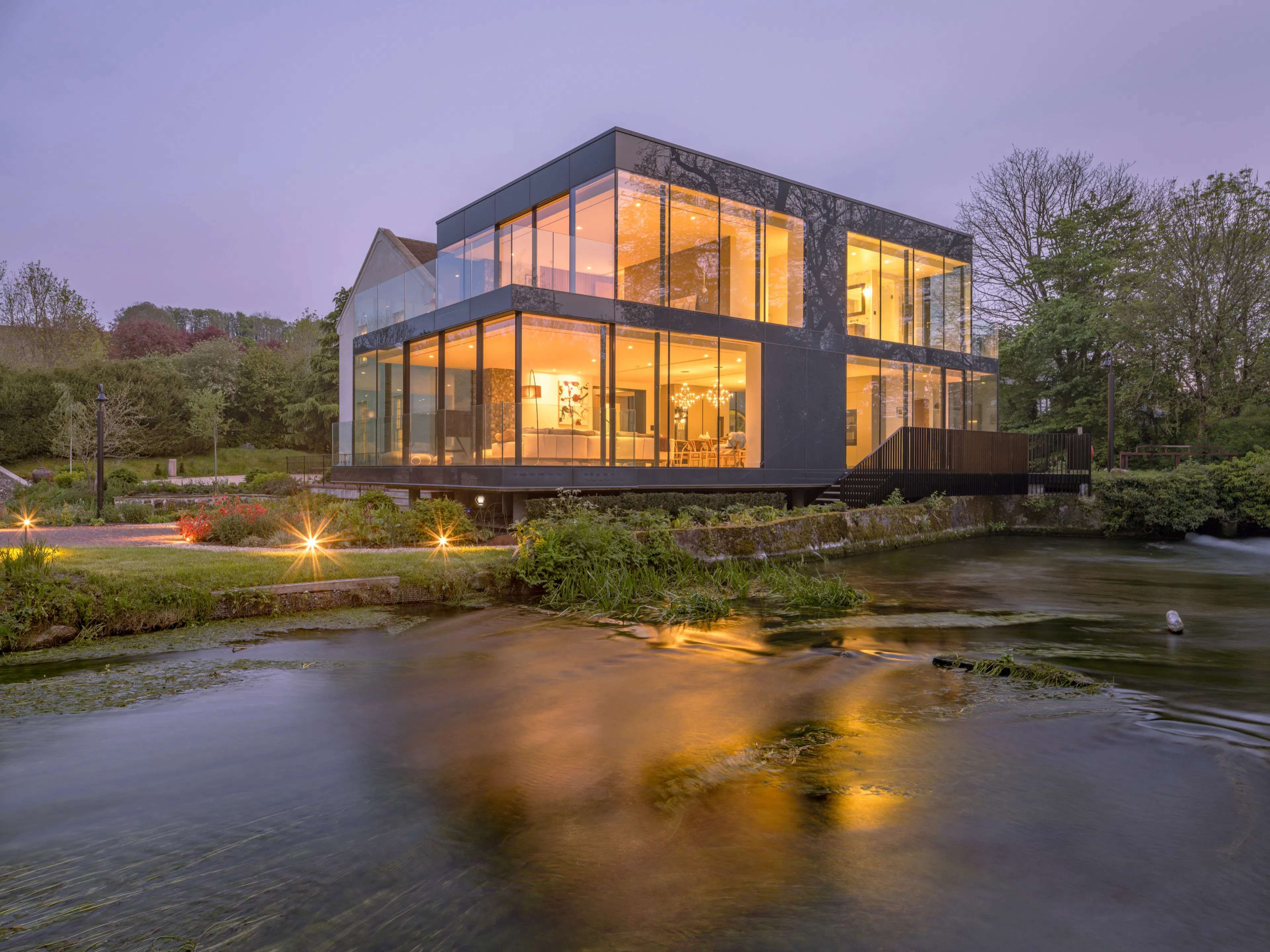 Glass wing of a Wiltshire mill house lit from within at dusk, reflected in the river below