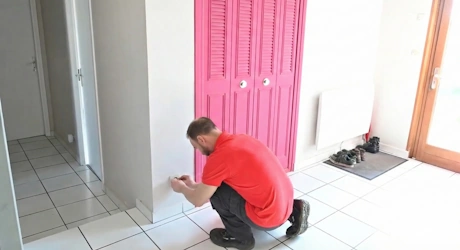 Technicien en rouge accroupi devant un radiateur rose dans une pièce carrelée