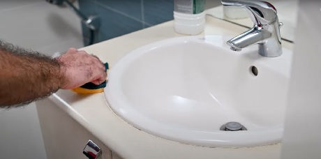 Main lissant le joint silicone neuf autour d'un lavabo avec une éponge