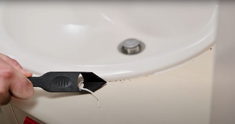 Main grattant l'ancien joint silicone autour d'un lavabo avec un outil noir