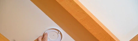 Main installant un détecteur de fumée au plafond près d'une poutre en bois