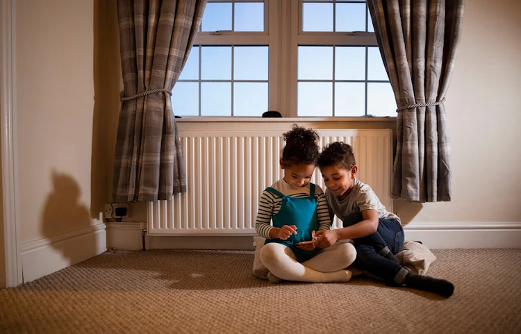 Deux enfants assis par terre devant un radiateur jouant avec une tablette