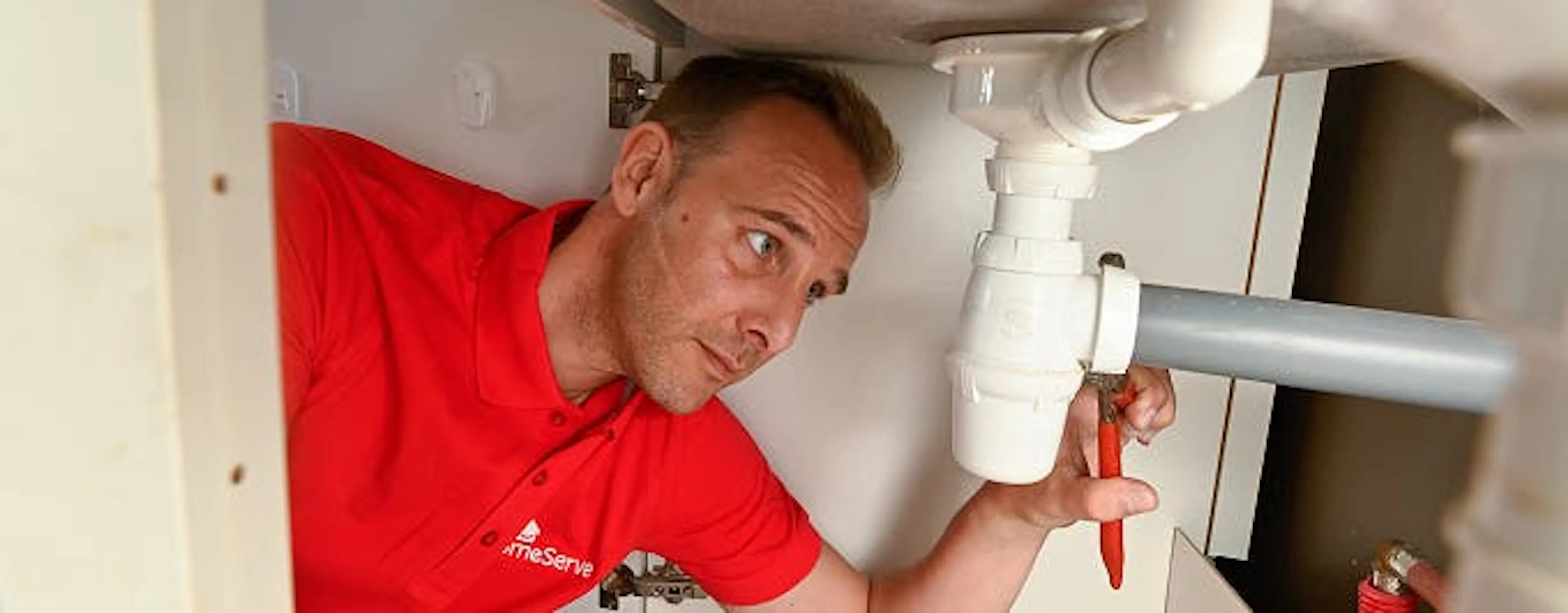 Plombier en polo rouge réparant la tuyauterie sous un évier