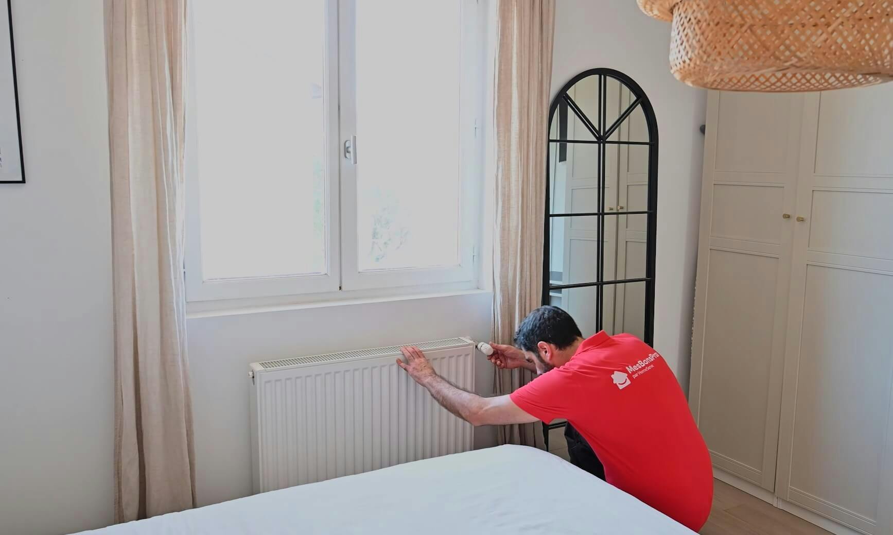 Technicien en rouge réparant un radiateur blanc sous une fenêtre