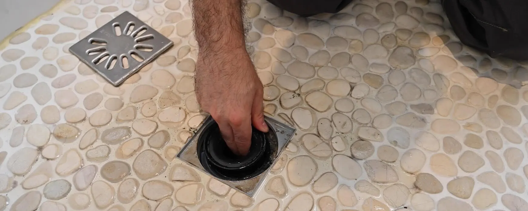Main en train de déboucher une bonde d'une douche sur carrelage en galets