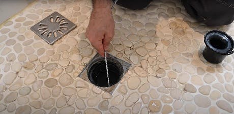 Main nettoyant la bonde d'une douche à galets avec un fil