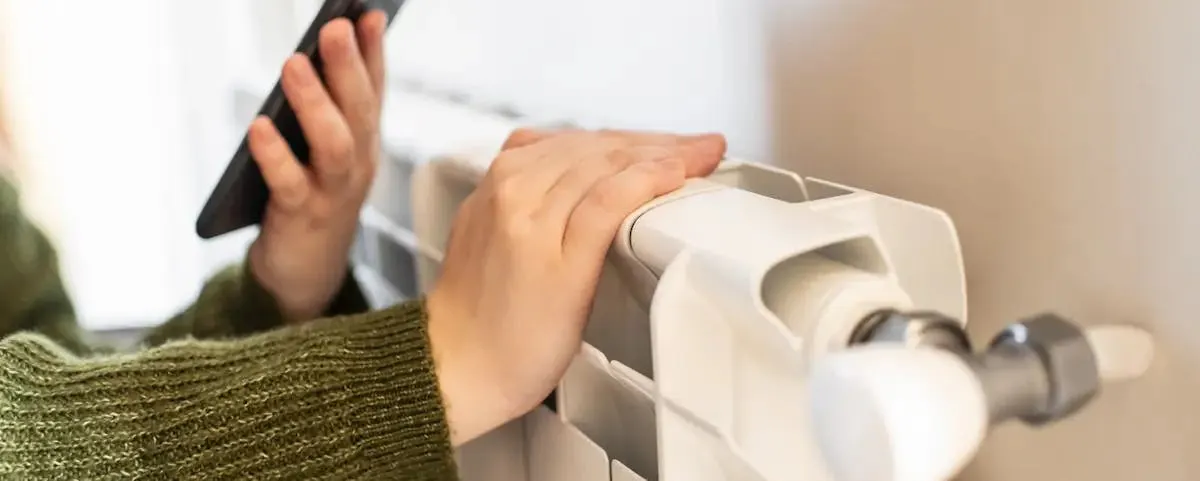 Main sur un radiateur blanc et smartphone pour calculer le coût d'un degré supplémentaire de chauffage