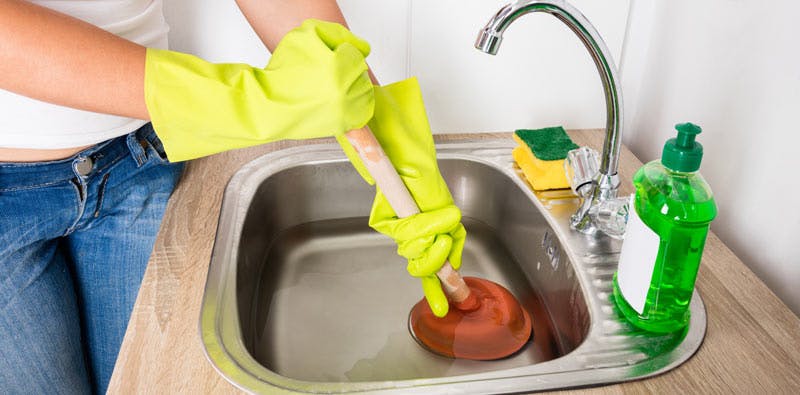 Personne portant des gants en caoutchouc jaunes utilisant une ventouse pour déboucher un évier de cuisine